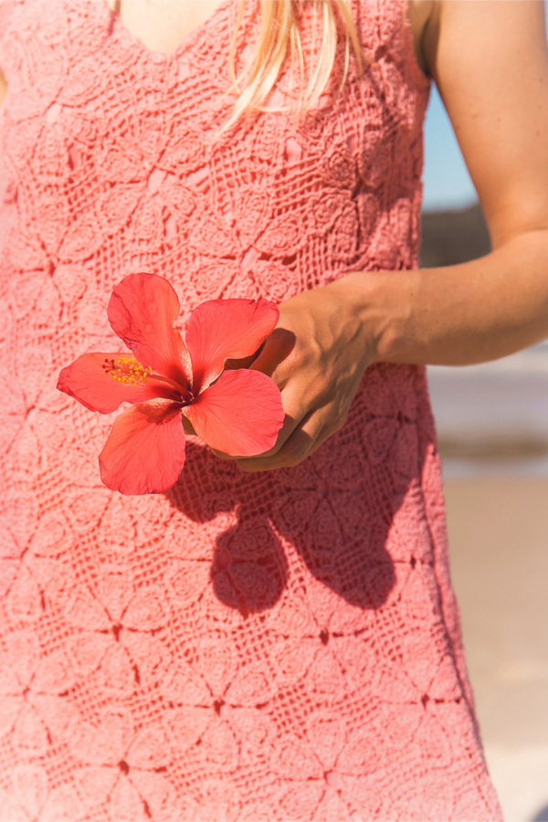 A close-up of a person wearing a coral pink crocheted sleeveless dress, holding a bright pink hibiscus flower against a sunny outdoor background.