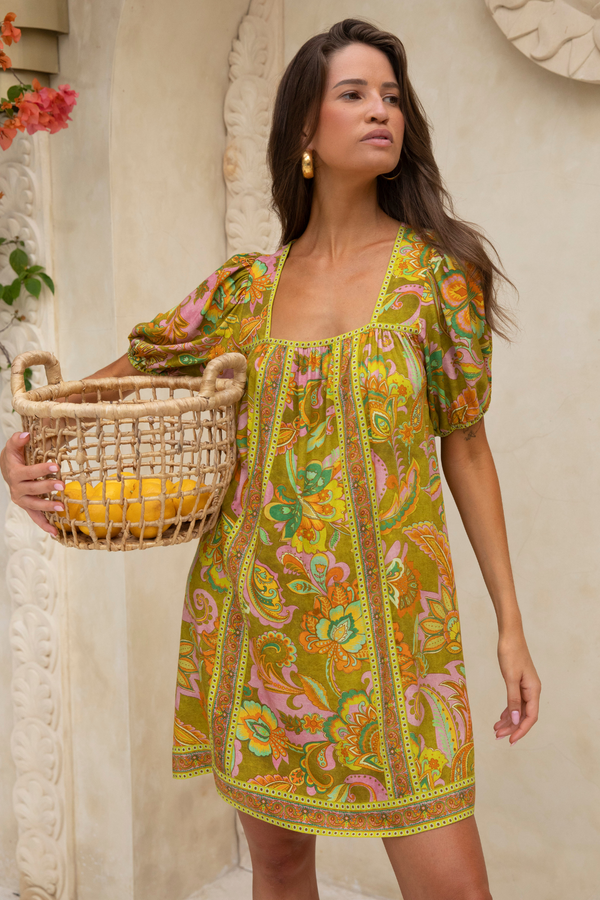 Woman wearing a colorful dress holding a woven basket against a neutral wall.