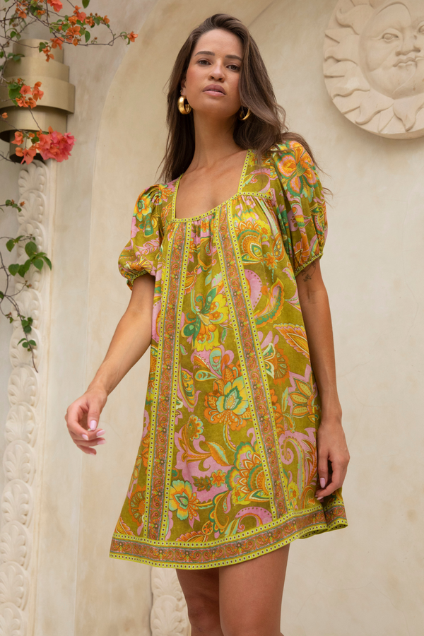 Woman wearing a colorful patterned dress standing against a decorative wall.