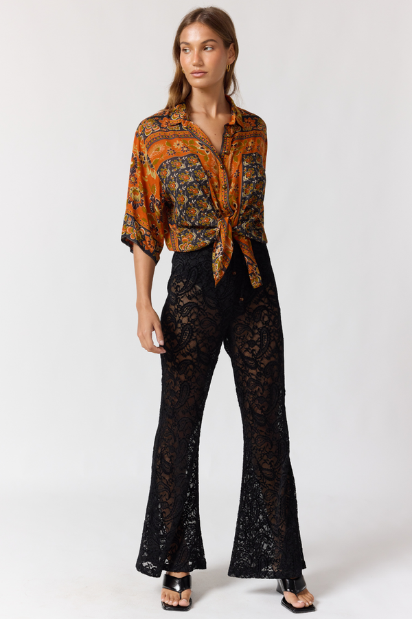 Woman wearing a patterned orange blouse and black lace pants on a white background