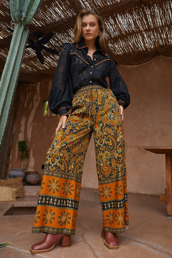Woman wearing a black shirt and patterned orange pants standing in a rustic setting with cacti and a thatched roof.