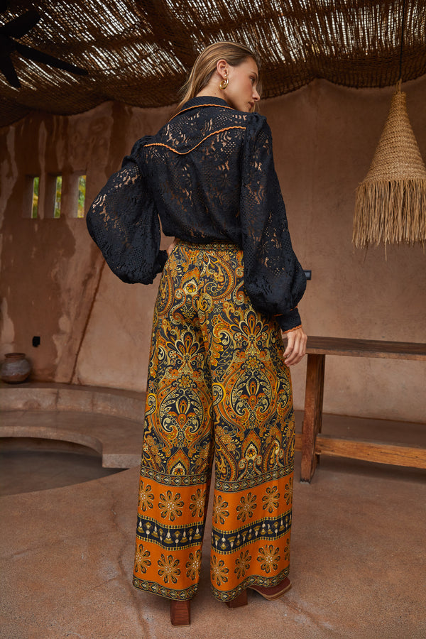 Woman wearing a dark blue embroidered top and patterned orange and yellow pants in a rustic setting.