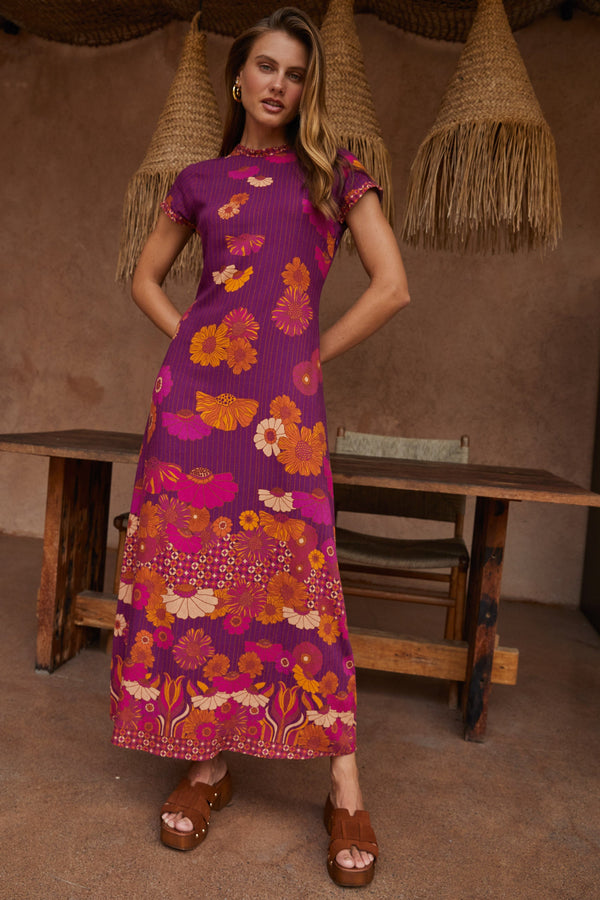 Woman wearing a purple floral dress standing in a rustic setting with wooden tables and straw hats.