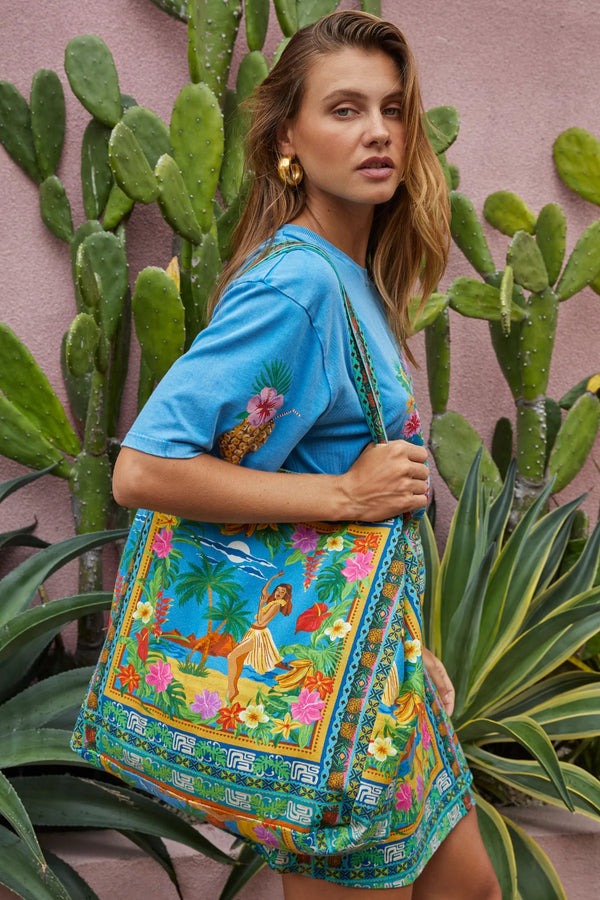 Woman holding a colorful handbag with a desert scene design against a pink wall with cacti.