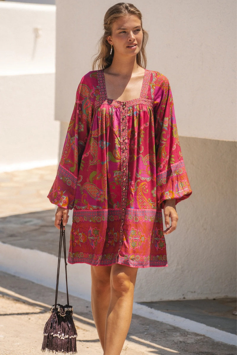 Woman wearing a pink floral dress holding a black handbag against a white wall.