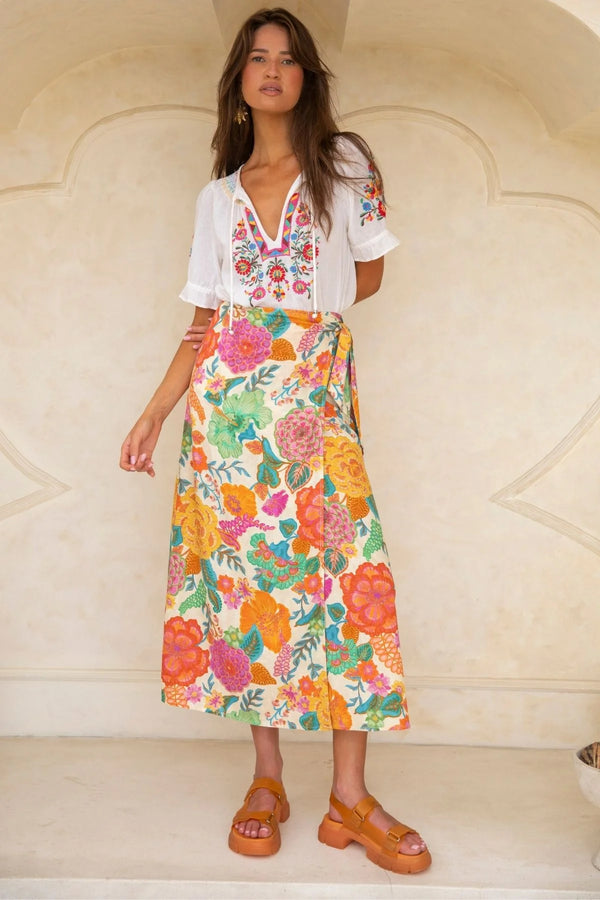 Woman wearing a white blouse with floral patterns and a colorful skirt against a beige wall.