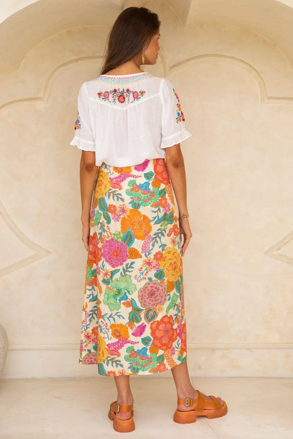 Woman wearing a white blouse with floral embroidery and a colorful floral skirt against a beige wall.