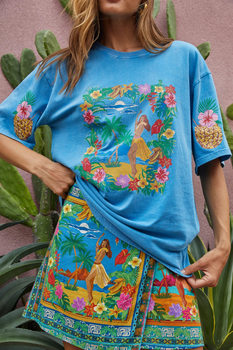 Person wearing a blue t-shirt and colorful skirt with tropical designs and pineapple graphics.