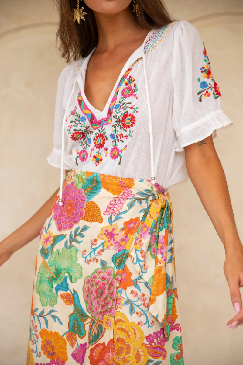 Woman wearing a white blouse with colorful floral embroidery and a matching colorful skirt against a beige background.