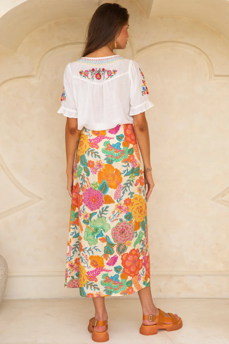 Woman wearing a white embroidered top and colorful floral skirt against a beige wall.