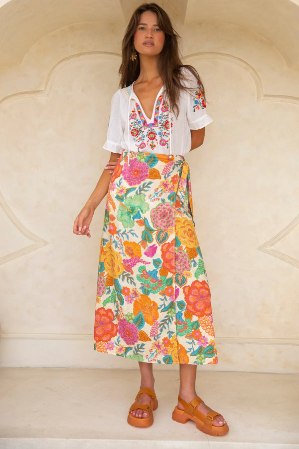 Woman wearing a white blouse with floral embroidery and a colorful floral skirt against a beige wall.