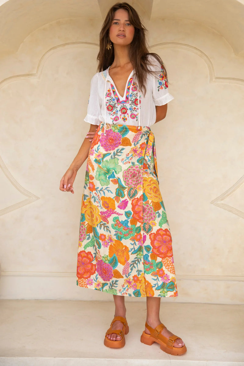 Woman wearing a white blouse with floral embroidery and a colorful floral skirt against a beige wall.