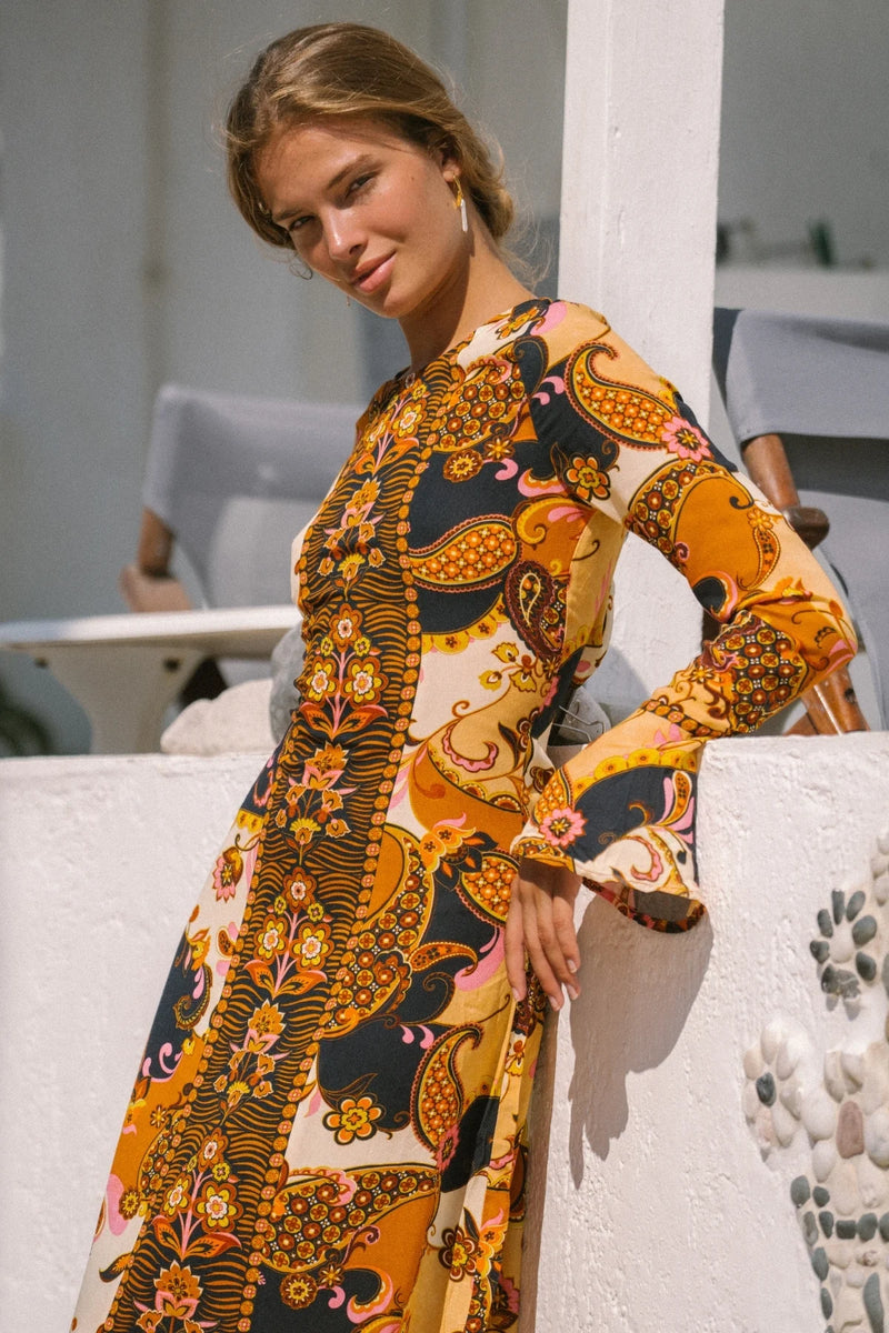 Woman wearing a patterned dress leaning against a white wall.