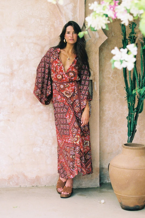 Woman in a patterned dress standing in a decorative setting with plants and a vase.