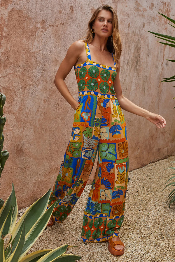 Woman wearing a colorful patterned jumpsuit standing against a textured wall with plants.