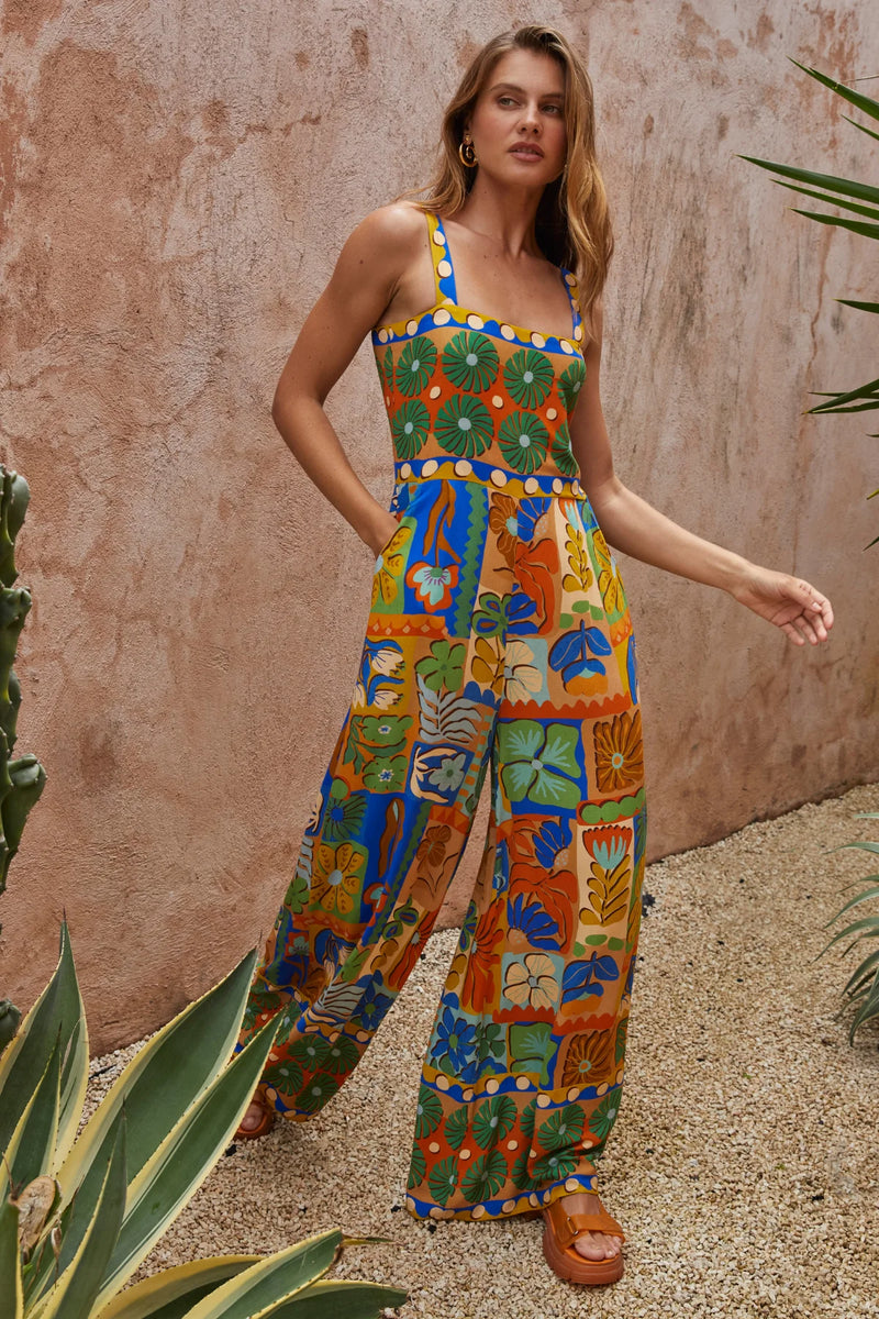 Woman wearing a colorful patterned jumpsuit standing against a textured wall with plants.