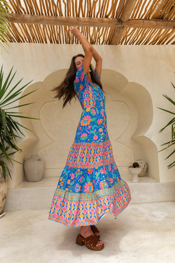 Woman in a colorful dress standing under a thatched roof with decorative wall and plants.