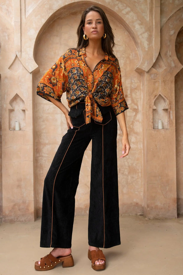 Woman wearing an orange patterned blouse and black pants standing in front of a beige architectural background.