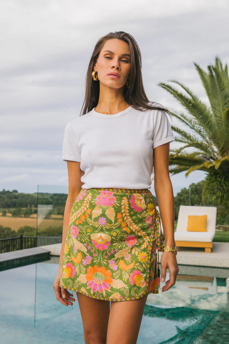 Woman wearing a white t-shirt and colorful floral skirt by a pool with palm trees in the background