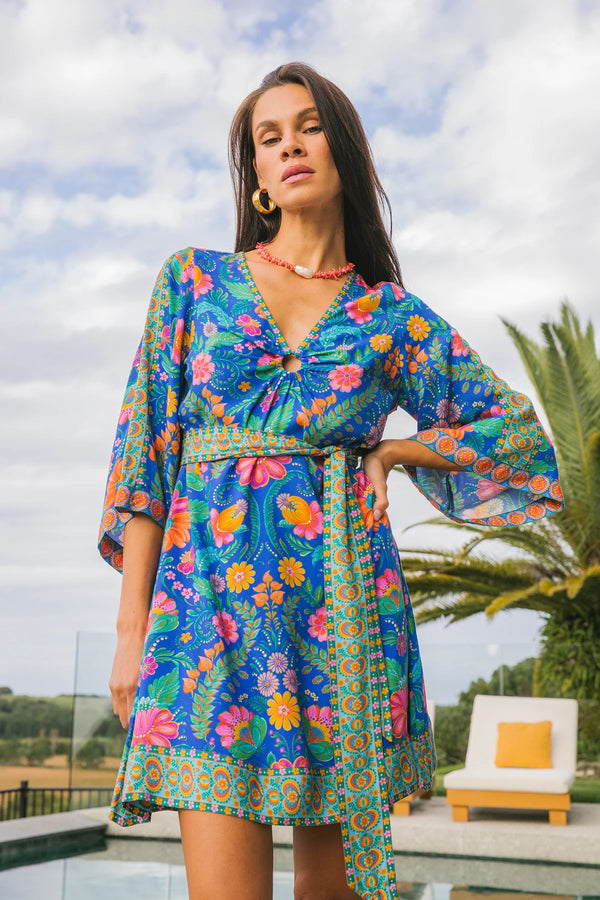 Woman wearing a colorful floral dress standing outdoors with palm trees and a cloudy sky in the background.