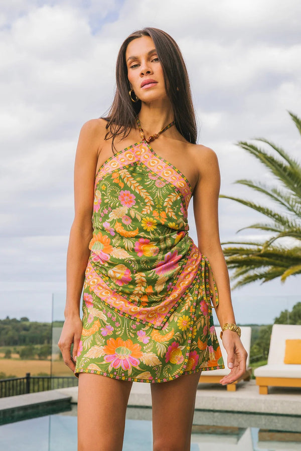 Woman wearing a colorful floral dress standing outdoors with palm trees and a pool in the background