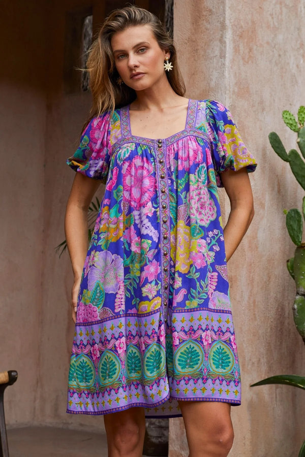 Woman wearing a colorful patterned dress standing against a rustic wall with a cactus plant.