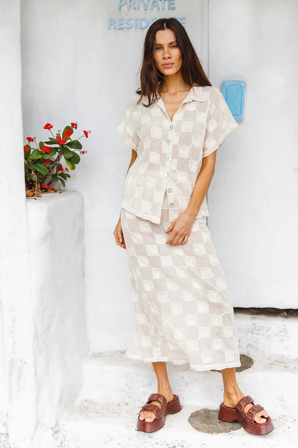 Woman wearing a white checkered outfit standing in front of a white wall with a plant and blue sign.