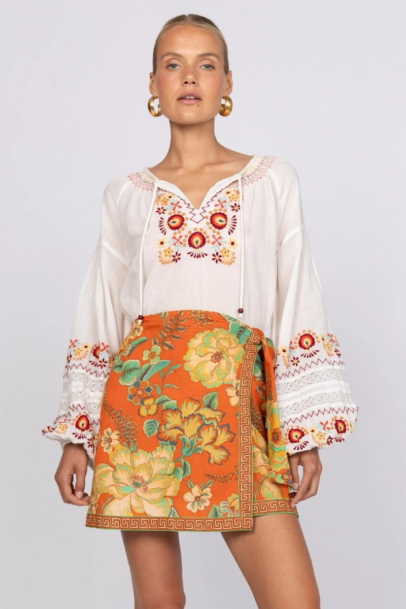 Woman wearing a white blouse with floral embroidery and an orange skirt with floral patterns on a plain background
