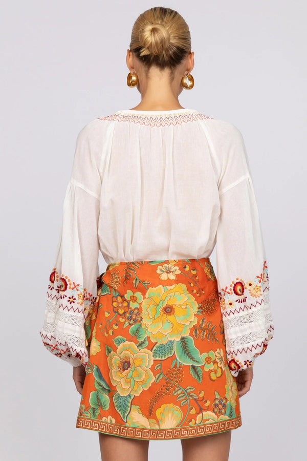 Person wearing a white blouse with floral embroidery and an orange skirt with similar design on a plain background
