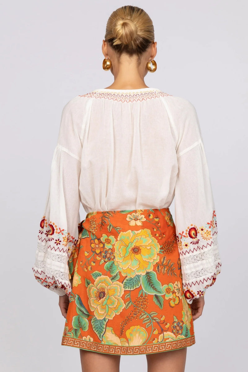 Person wearing a white blouse with floral embroidery and an orange skirt with similar design on a plain background