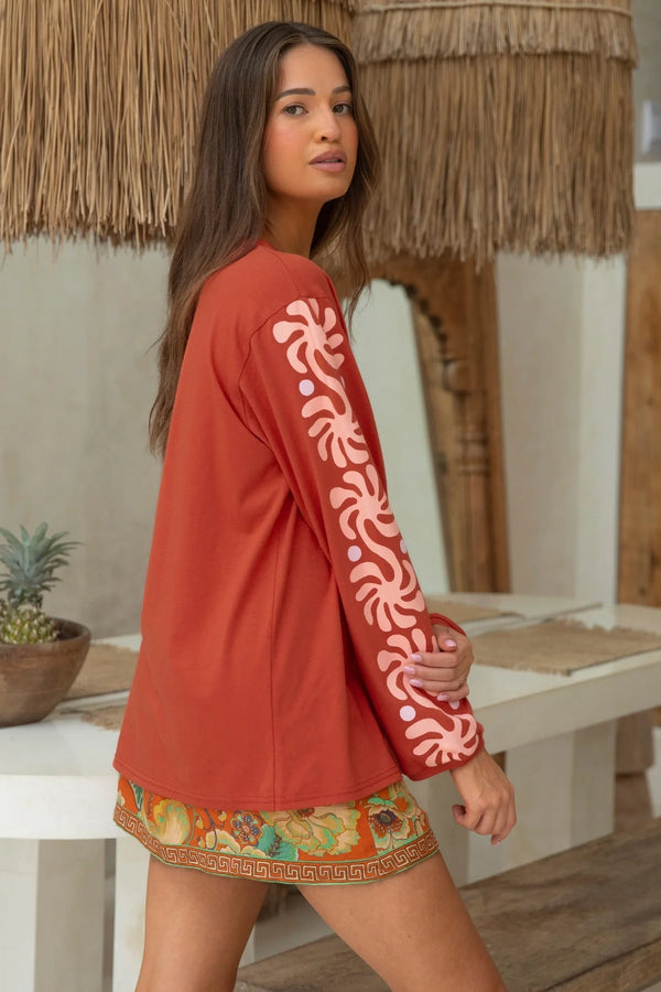 Woman wearing a red long-sleeve top with floral patterns and a colorful skirt in a tropical setting.