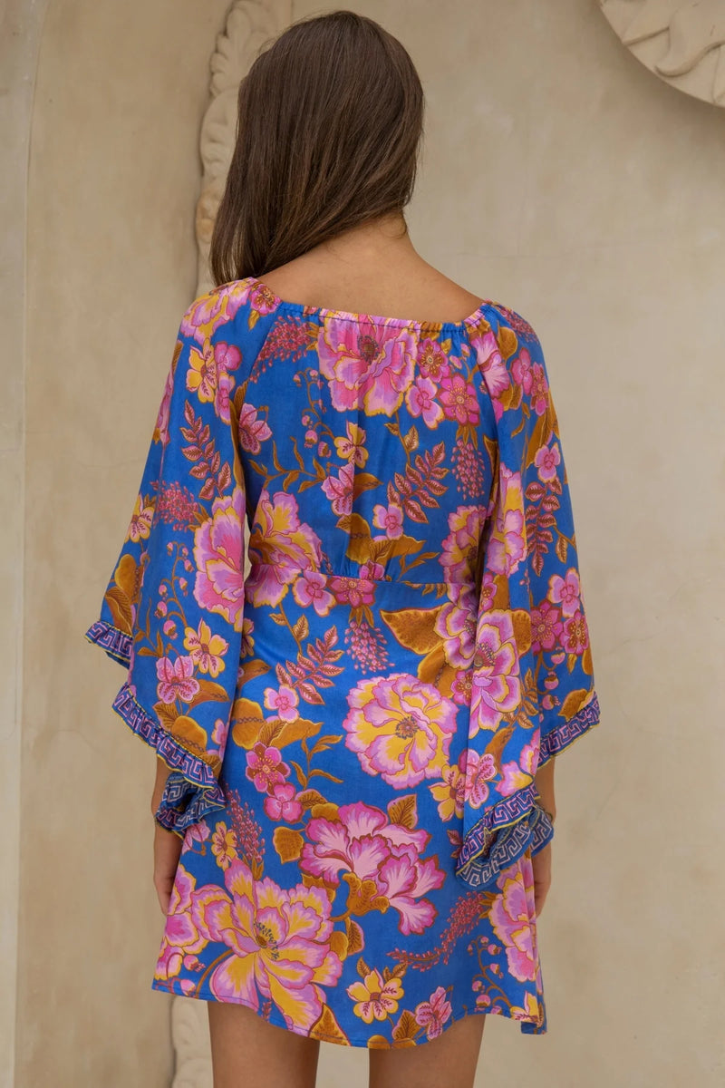 Woman wearing a blue floral dress with pink and orange flowers against a beige wall.