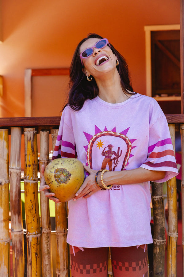 A woman wearing a light purple oversized graphic t-shirt with pink and white striped sleeves, holding a large yellow coconut in front of a bamboo fence and orange wall.