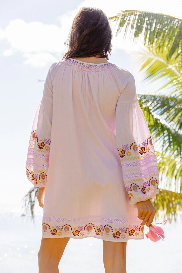 A woman viewed from behind wearing a light pink bohemian-style tunic dress with embroidered floral details on the sleeves and hem, set against a tropical beach background with palm trees.
