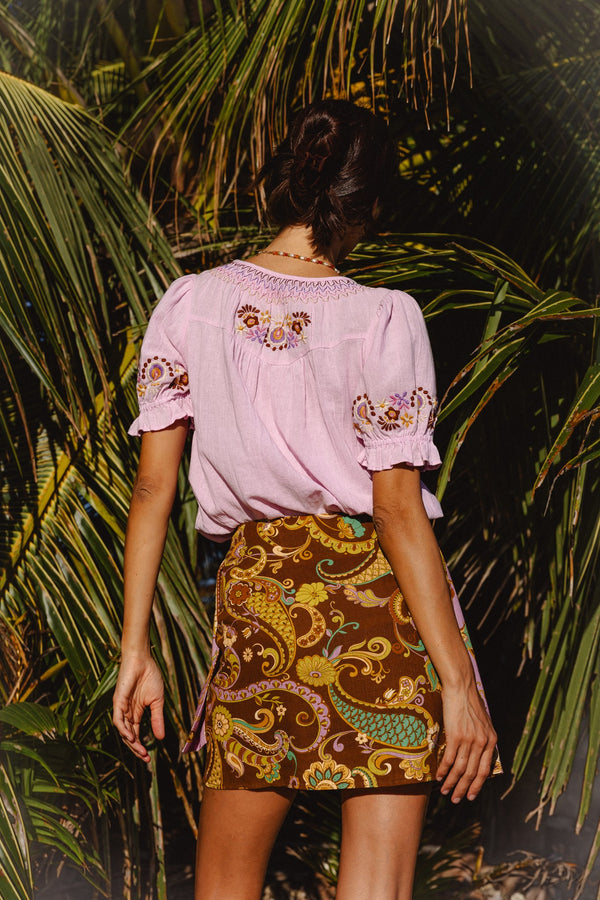 A woman viewed from behind wearing a light pink embroidered blouse and a brown patterned mini skirt, standing outdoors in front of palm leaves.