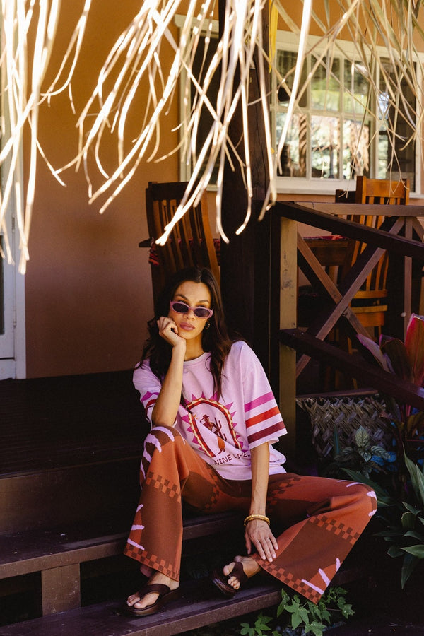 A woman sits on dark wooden steps of a tropical resort, wearing a light pink t-shirt with a red graphic print and matching brown patterned wide-leg pants.