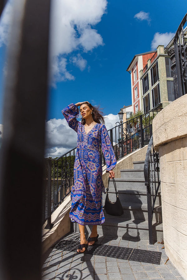 Woman in a blue patterned dress standing on a stone staircase with buildings and blue sky in the background.