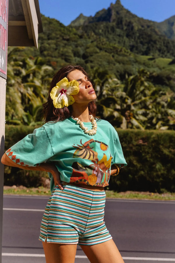 A woman posing outdoors in a vibrant mint green crop top with a colorful tropical print and matching striped shorts, set against a lush tropical mountain background. Location is Cook Islands.