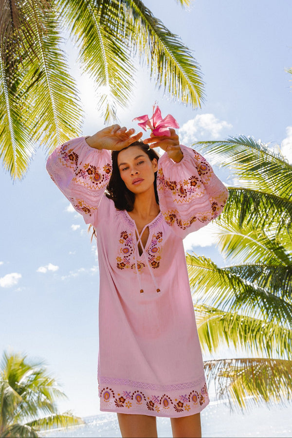 A woman wearing a light pink embroidered beach cover-up dress stands outdoors under palm trees against a bright blue sky, holding a pink flower.