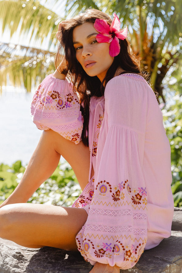 A woman sitting outdoors on a rock wearing a light pink bohemian-style embroidered cover-up dress with long puff sleeves and a pink flower in her hair.