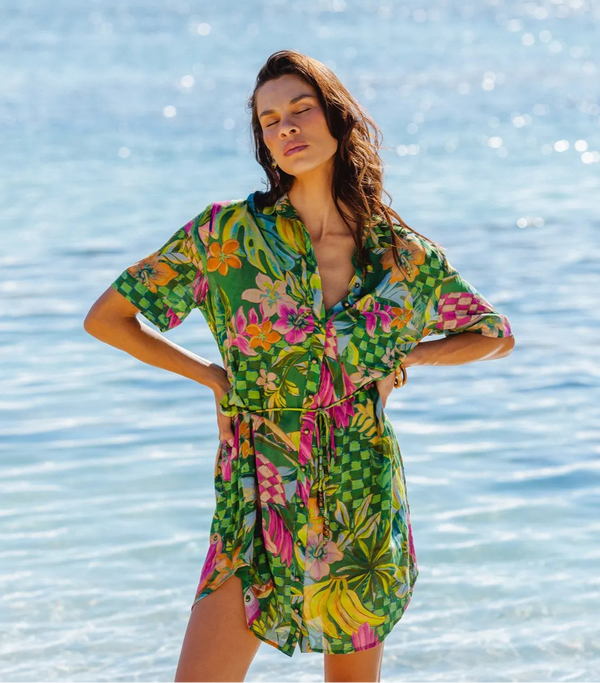 A woman stands in shallow ocean water wearing a short-sleeve tropical print shirt dress. The dress has a green base with pink, yellow, and orange floral and leaf patterns, and a tie-front waist.
