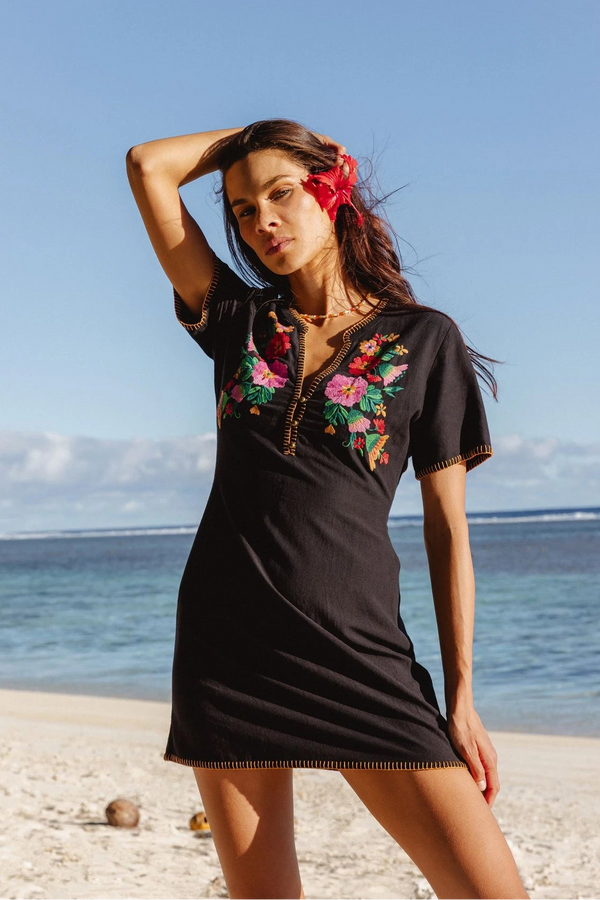Woman in a black dress with floral embroidery standing on a beach.
