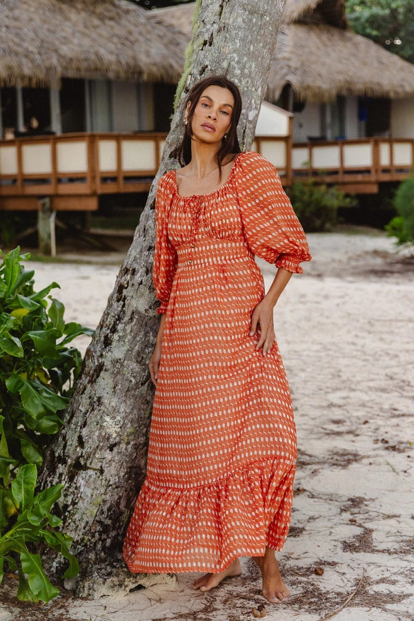 A woman wearing a rust-orange and white patterned maxi dress leans against a tree on a sandy beach, with thatched-roof bungalows in the background.