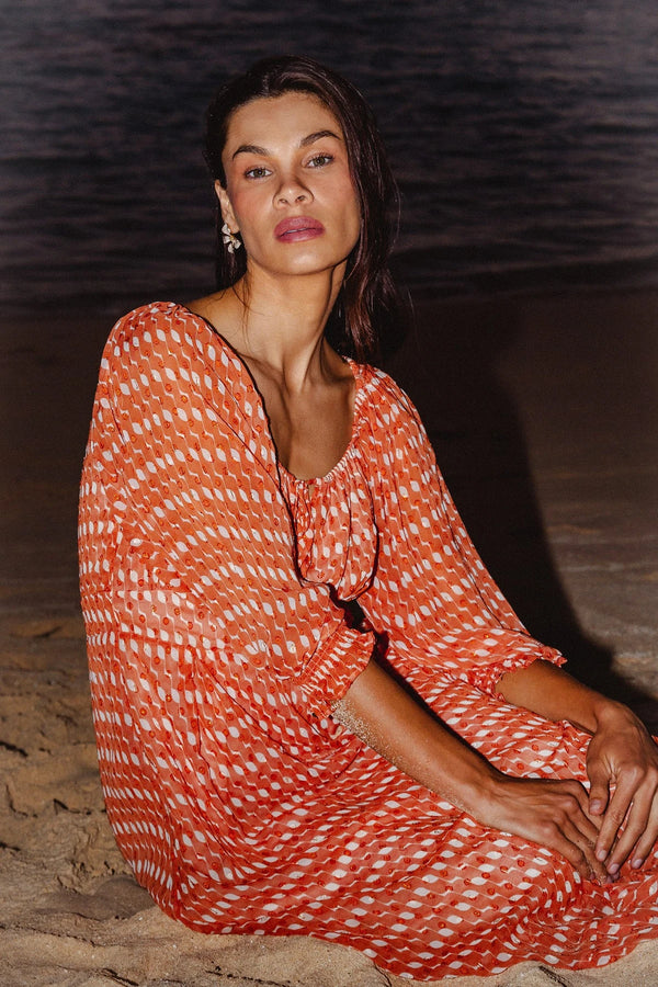 A woman sitting on a sandy beach at night wearing a loose, long-sleeve orange and white patterned dress.