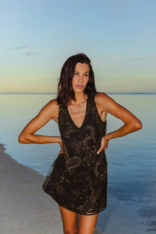 A woman stands on a beach wearing a sleeveless black lace dress with a shimmering gold abstract pattern. The background features calm water under a soft blue sky.