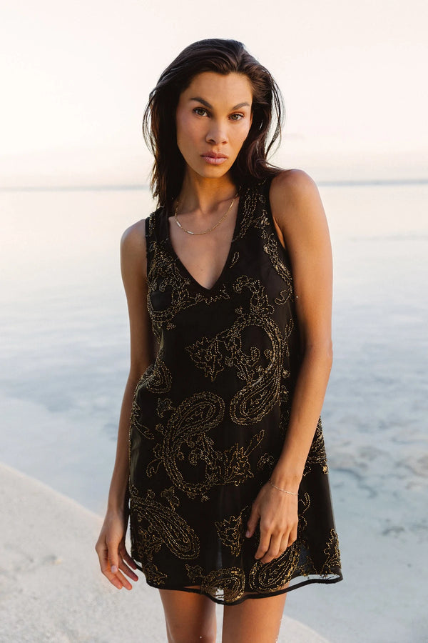 A woman wearing a sleeveless black dress with a gold paisley pattern, standing on a beach with the ocean in the background.