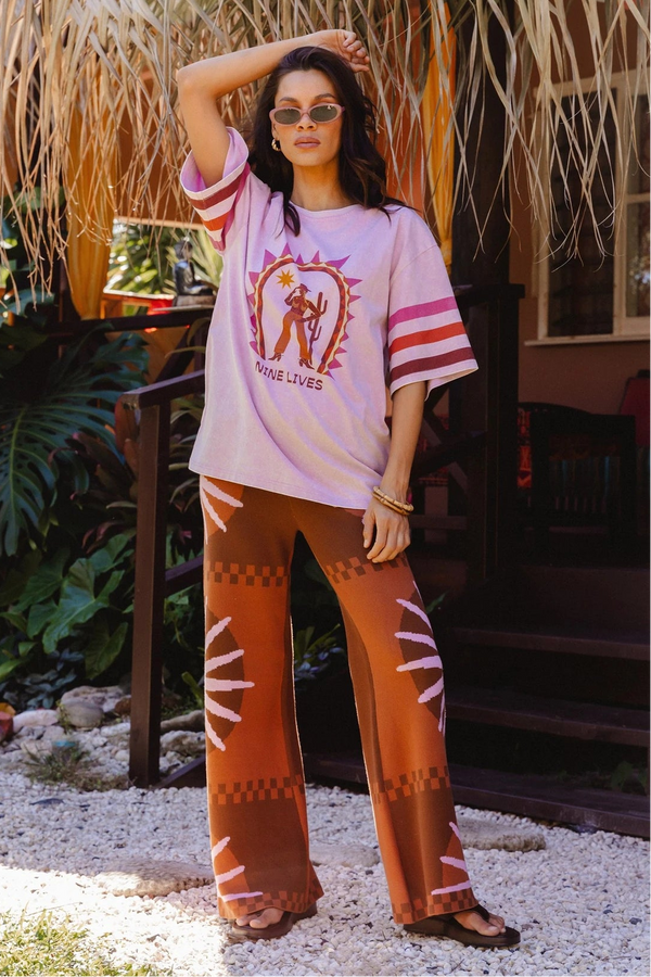 A woman stands outdoors in front of a thatched roof and tropical plants, wearing a light pink graphic T-shirt with pink and white striped sleeves and brown wide-leg pants with white geometric patterns.