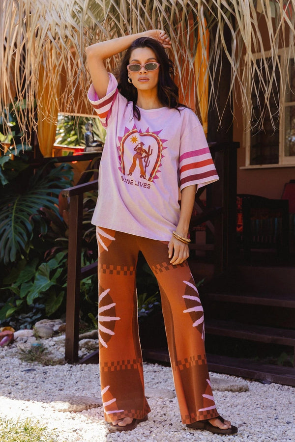 A woman stands outdoors in front of a thatched roof and tropical plants, wearing a light pink graphic T-shirt with pink and white striped sleeves and brown wide-leg pants with white geometric patterns.