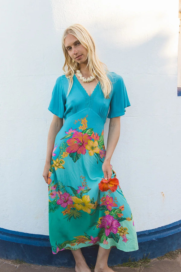 A woman wearing a turquoise short-sleeve midi dress with a vibrant multicolor floral print, standing barefoot against a white stucco wall.