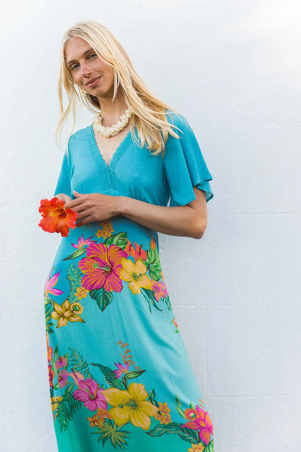 A woman wearing a turquoise blue maxi dress with a vibrant pink, yellow, and green tropical floral print, holding a red hibiscus flower against a white wall.